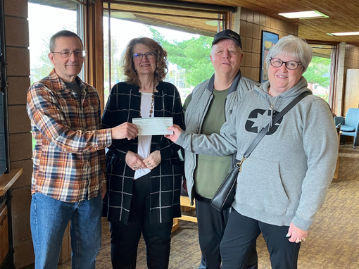 Four adults pose indoors while holding a check in front of large windows.